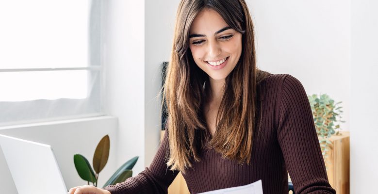 Young happy woman teleworking from home office. Professional female working using laptop computer while reading business documents sitting on table at the office. Teleworking concept.